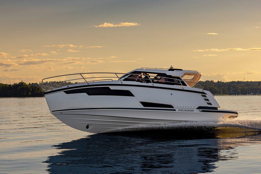 A sleek white boat named Aquador glides over calm water at sunset, with trees and clouds in the background.